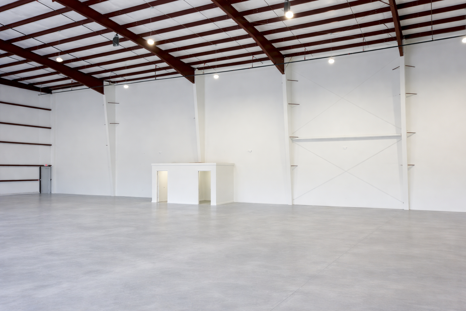 Empty warehouse or industrial space with white walls, high ceiling with metal framing, and a small white booth or office area in the corner.