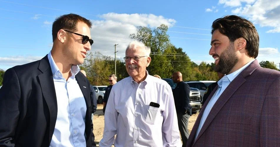 Three men are standing outdoors, engaged in conversation. Two of them are wearing suits, and the third is in a light-colored shirt. There are cars and trees in the background under a partly cloudy sky.