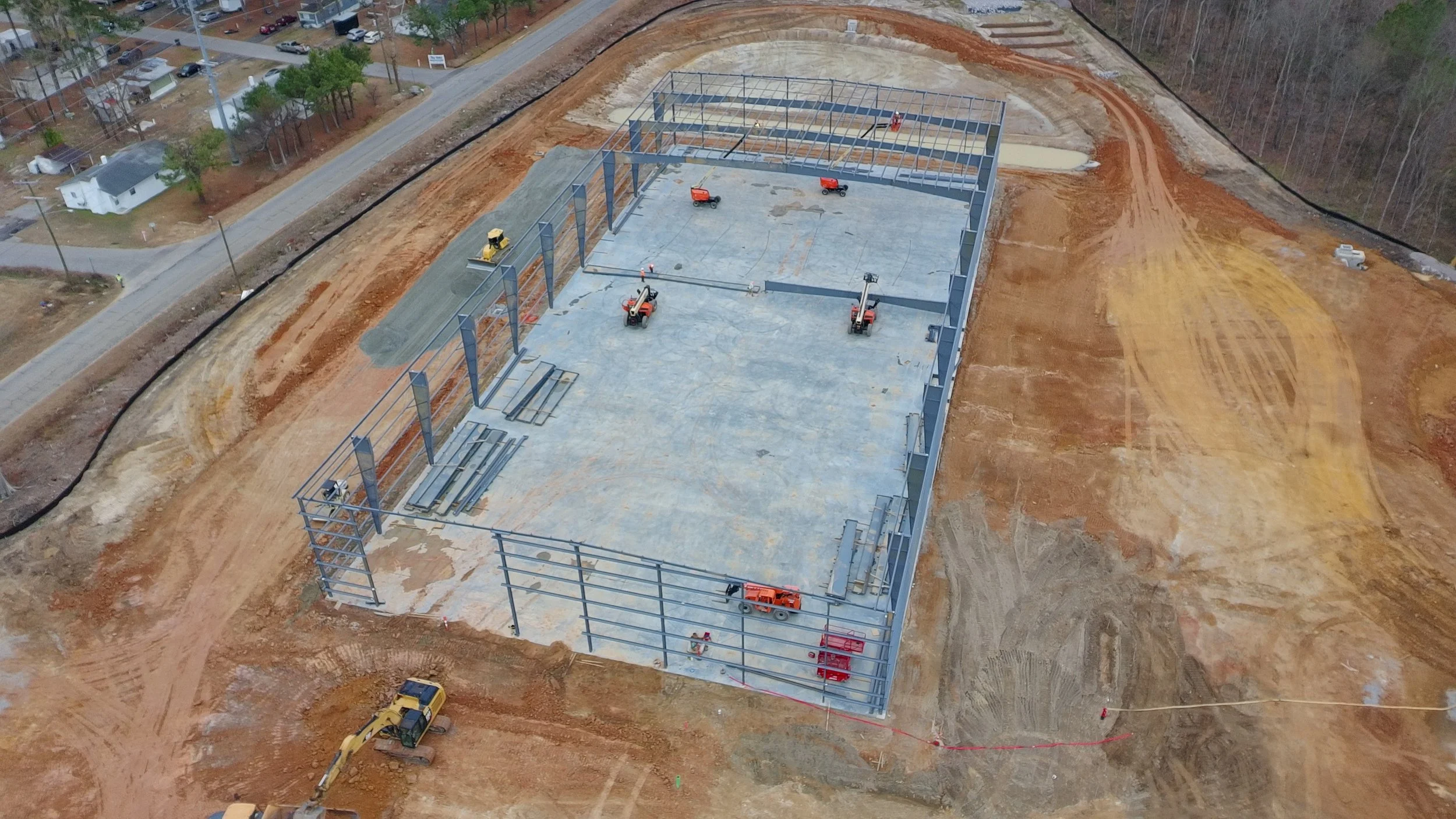 An aerial view of a construction site with a steel framework building being assembled on a concrete slab. Heavy machinery and construction materials are present on the site, with a surrounding dirt area and nearby residential neighborhood.