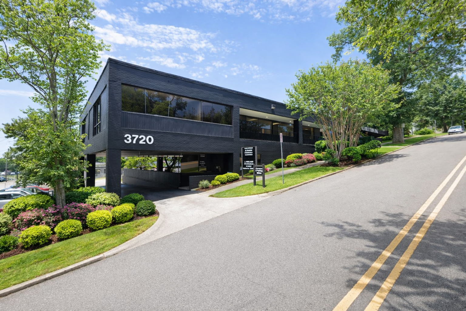 Black commercial building with the number 3720, surrounded by landscaped bushes and trees, on a sloped street under a partly cloudy sky.