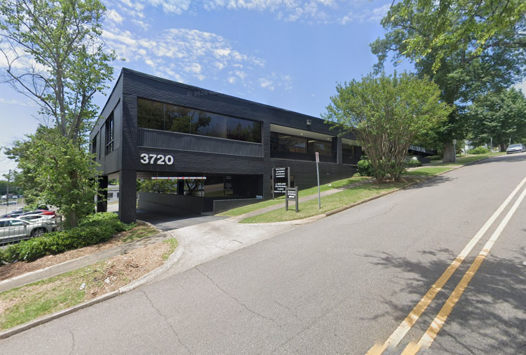 Black commercial building with the address 3720 on it, situated on a sloped street with trees and parking lot nearby, under a partly cloudy sky.