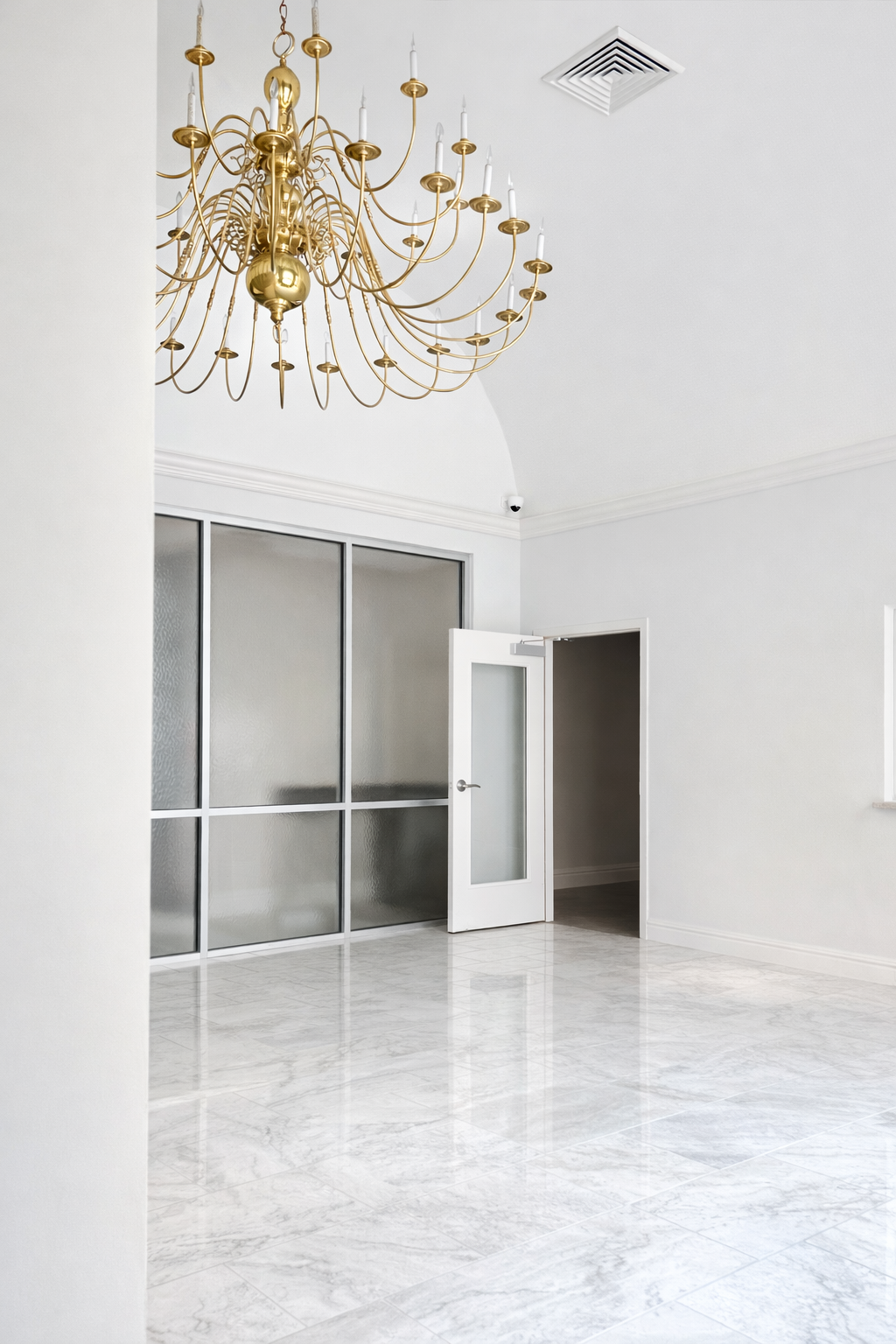 Empty white room with a large gold chandelier hanging from the ceiling, a half-open door, and frosted glass windows.