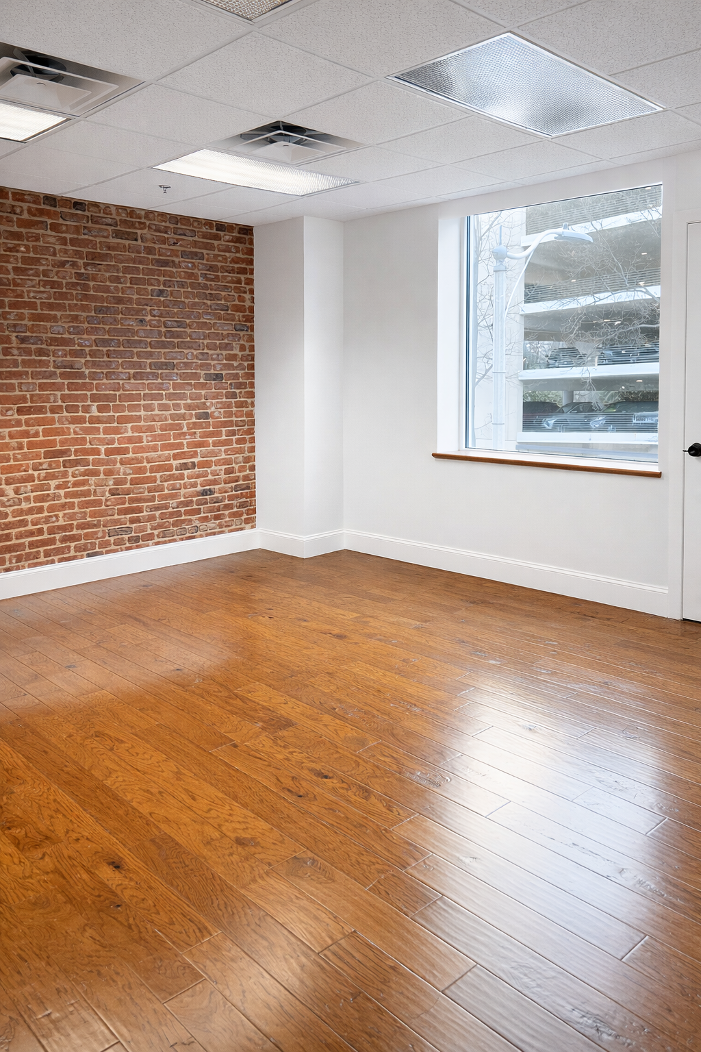 Empty room with hardwood floors, a brick accent wall, white walls, and a large window with a view of a parking garage.