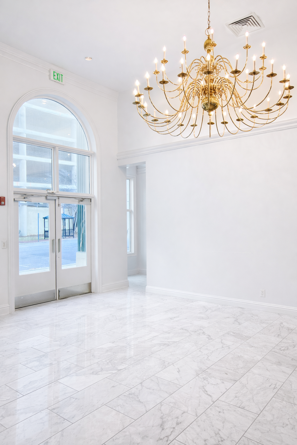 Empty white room with marble flooring, large arched window, and a gold chandelier hanging from the ceiling.