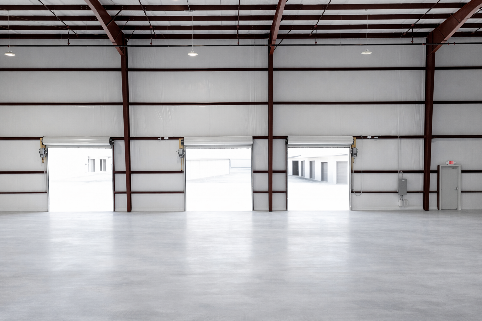 Empty warehouse with three open garage doors letting in bright natural light, white walls, concrete floor, exposed metal beams, and electrical wiring.