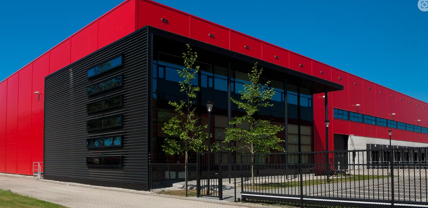 Modern red and black commercial building with large glass windows, trees, and a fenced walkway.
