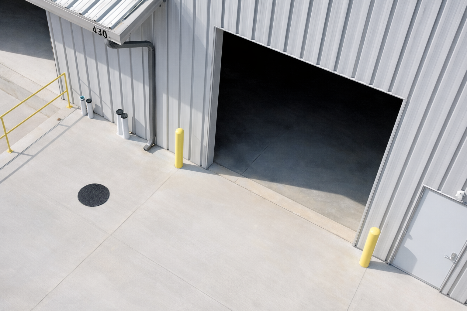 A large metal warehouse or storage building with an open garage door, a gray door with a handle, yellow and black safety bollards, pipes, and a yellow railing on a concrete floor.