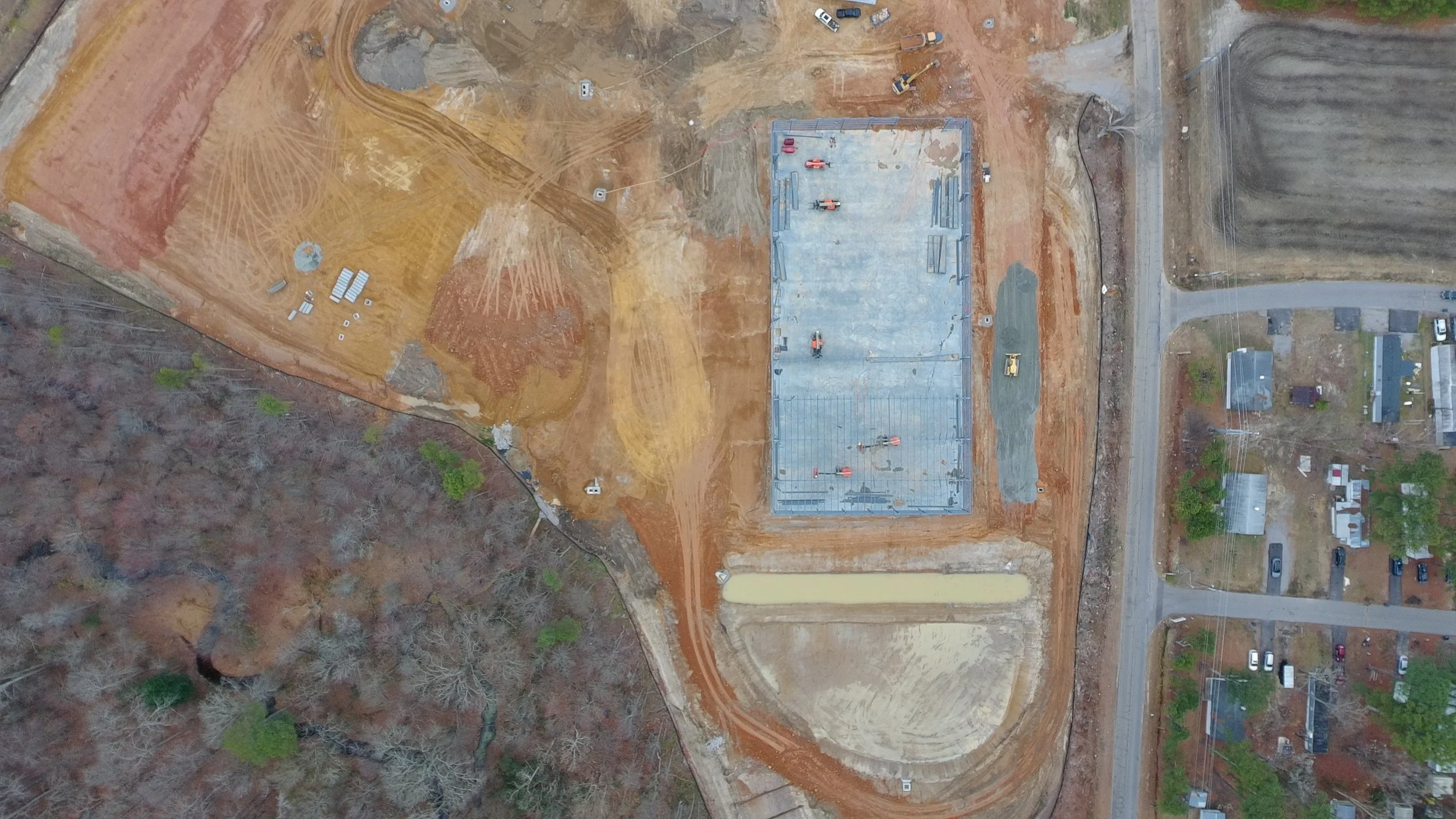 Drone view of a construction site with a rectangular building foundation, surrounding earthwork, and nearby roads and residential houses.
