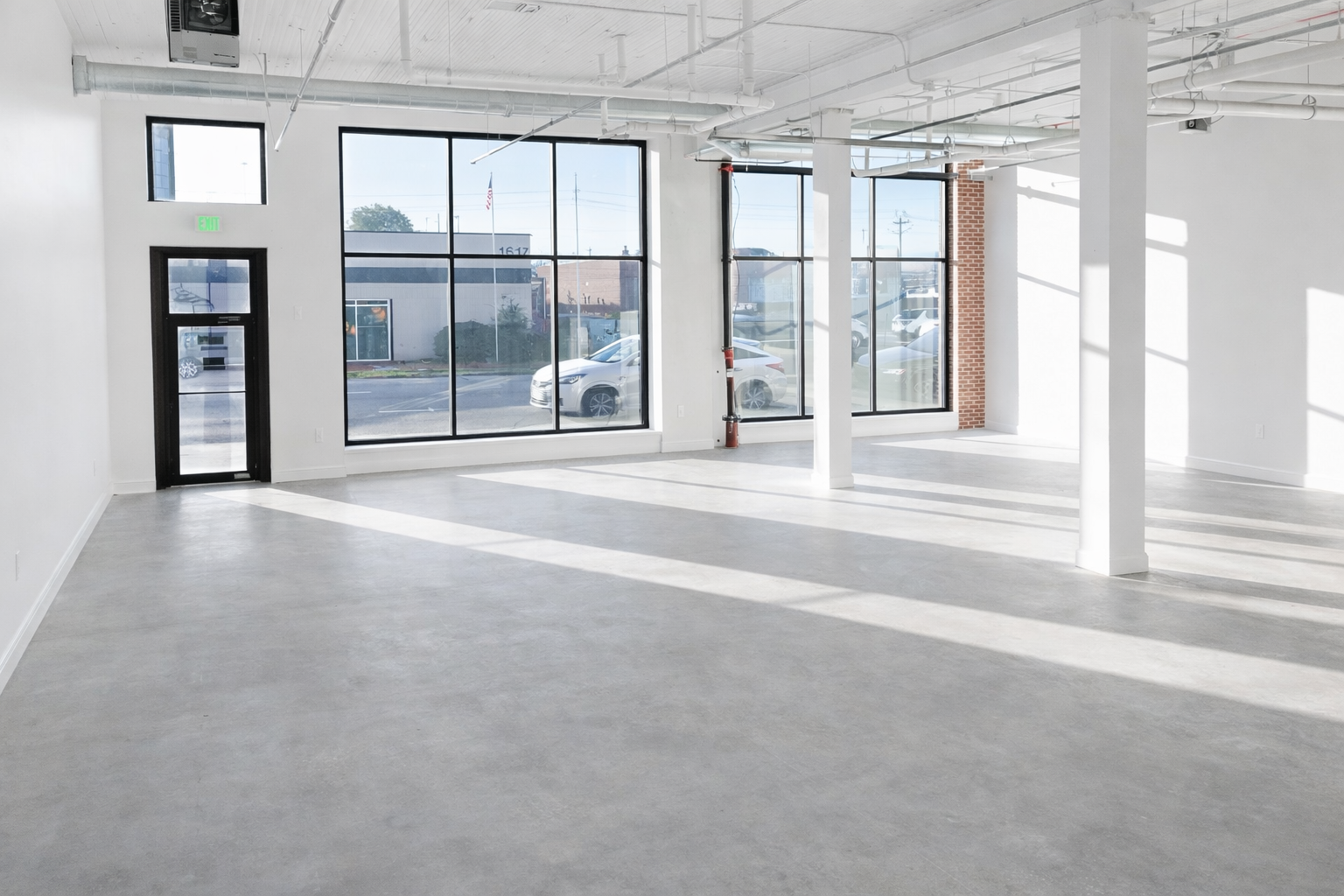 Empty commercial space with large windows, white walls, concrete floors, exposed ceiling pipes, and sunlight casting shadows.