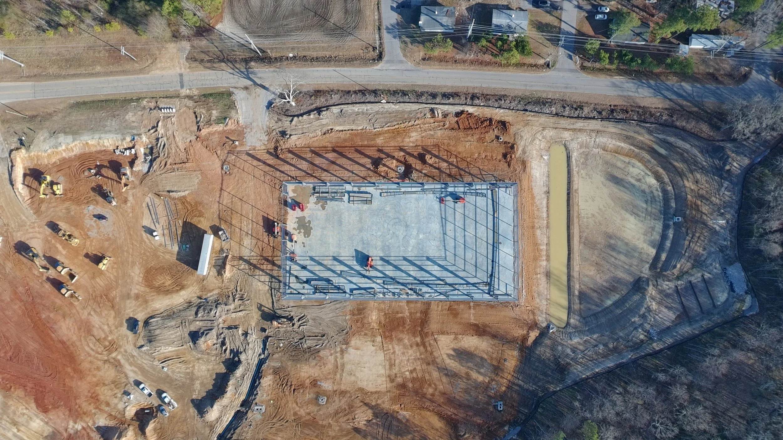 An aerial view of a construction site featuring a rectangular building foundation with steel framing, surrounded by dirt, construction equipment, and a few parked vehicles. A small concrete pond and a circular gravel area are also visible.