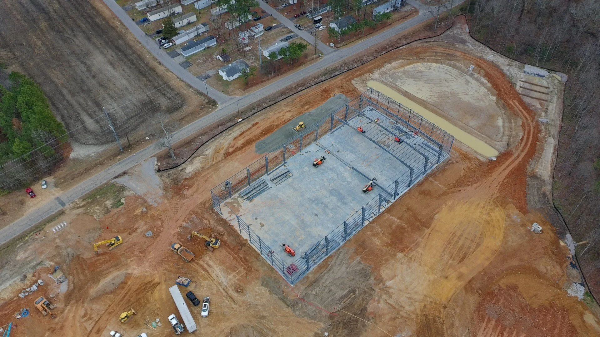 Aerial view of a construction site with a partially built sports facility foundation, surrounded by dirt roads, construction vehicles, and nearby residential houses and wooded area.