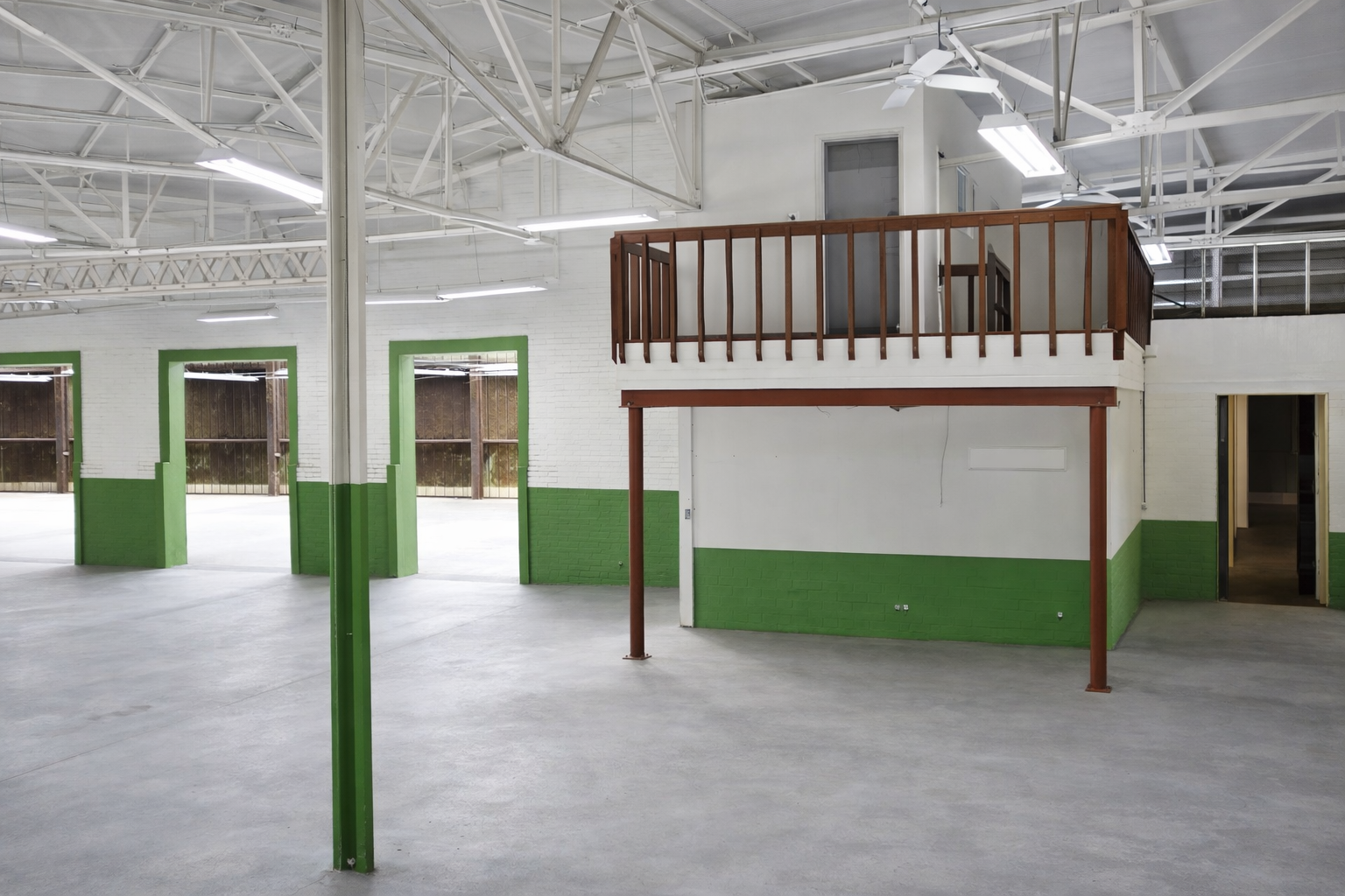 Interior view of a large, empty industrial or commercial space with white brick walls and green accents on the lower part of the walls. There are multiple windows, a mezzanine level with a wooden railing, and exposed ceiling beams with lighting fixtu
