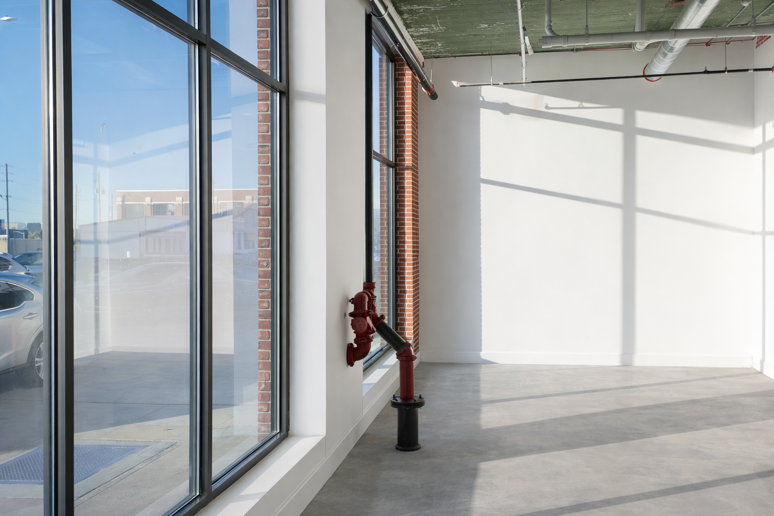 Empty commercial space with large windows, exposed ductwork, a red fire hydrant, and brick exterior visible through the window.