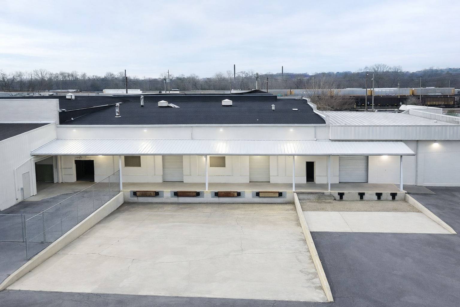 Empty parking lot in front of a commercial building with white walls and metal roofs, with train tracks and trees in the background under a cloudy sky.