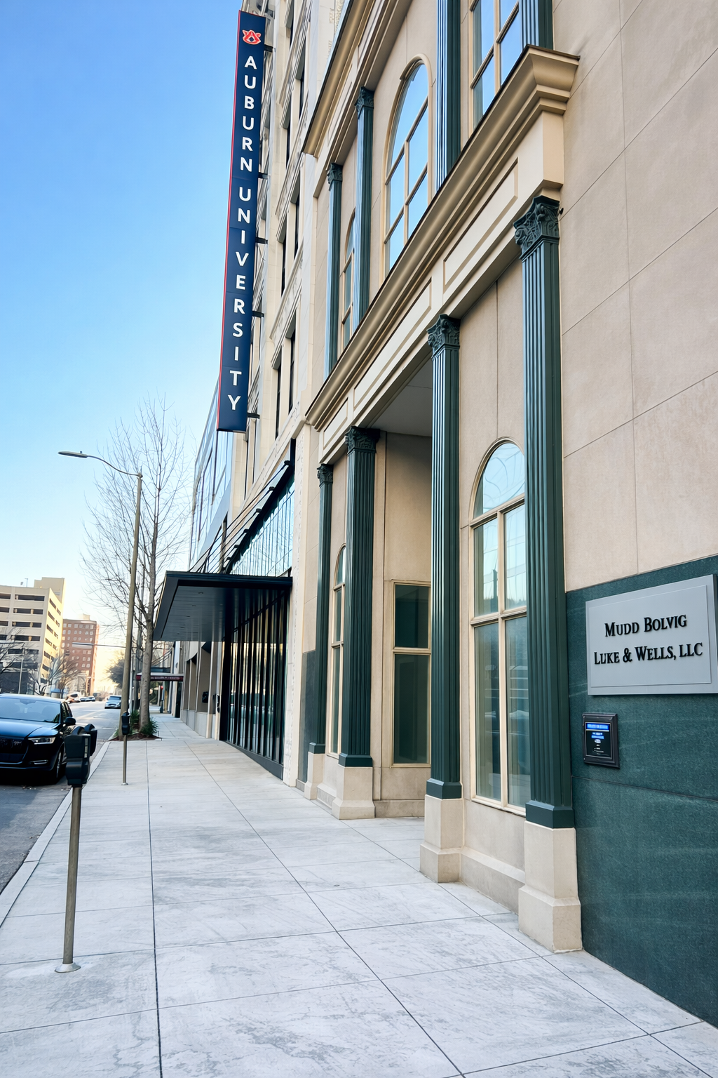 Sidewalk view of a modern building with a vertical sign for Auburn University, blue sky, and a street with parked cars.