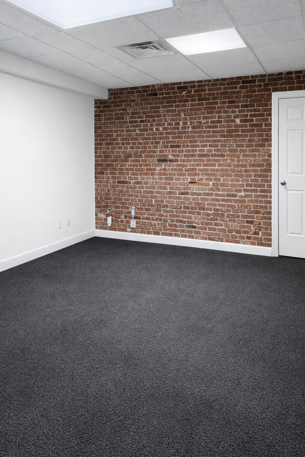 Empty room with a brick wall on one side, white wall on another, gray carpeted floor, drop ceiling with fluorescent lighting, and a closed white door.