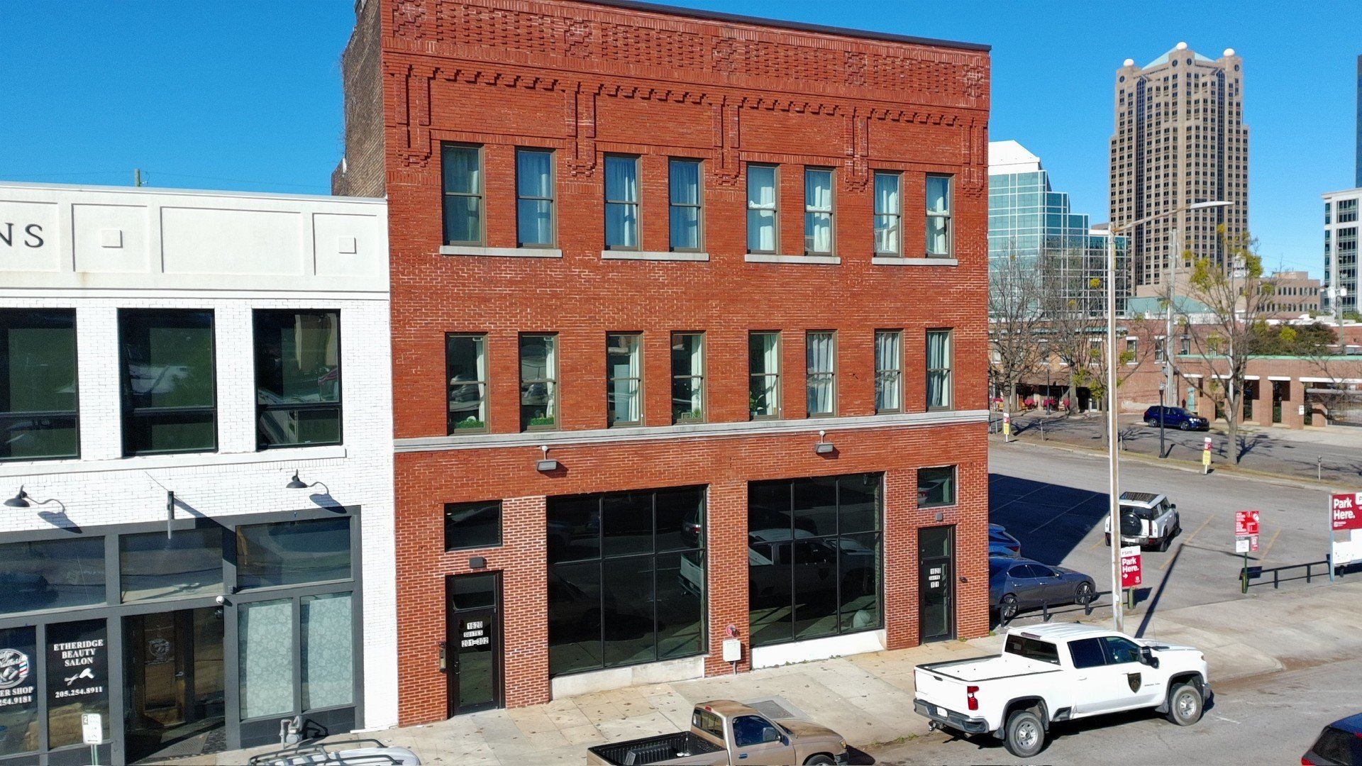 A brick building with large ground-floor glass windows and smaller upper-floor windows on a city street; neighboring white building with sign for a beauty salon; parked cars and a white pickup truck in front; city skyline in the background.