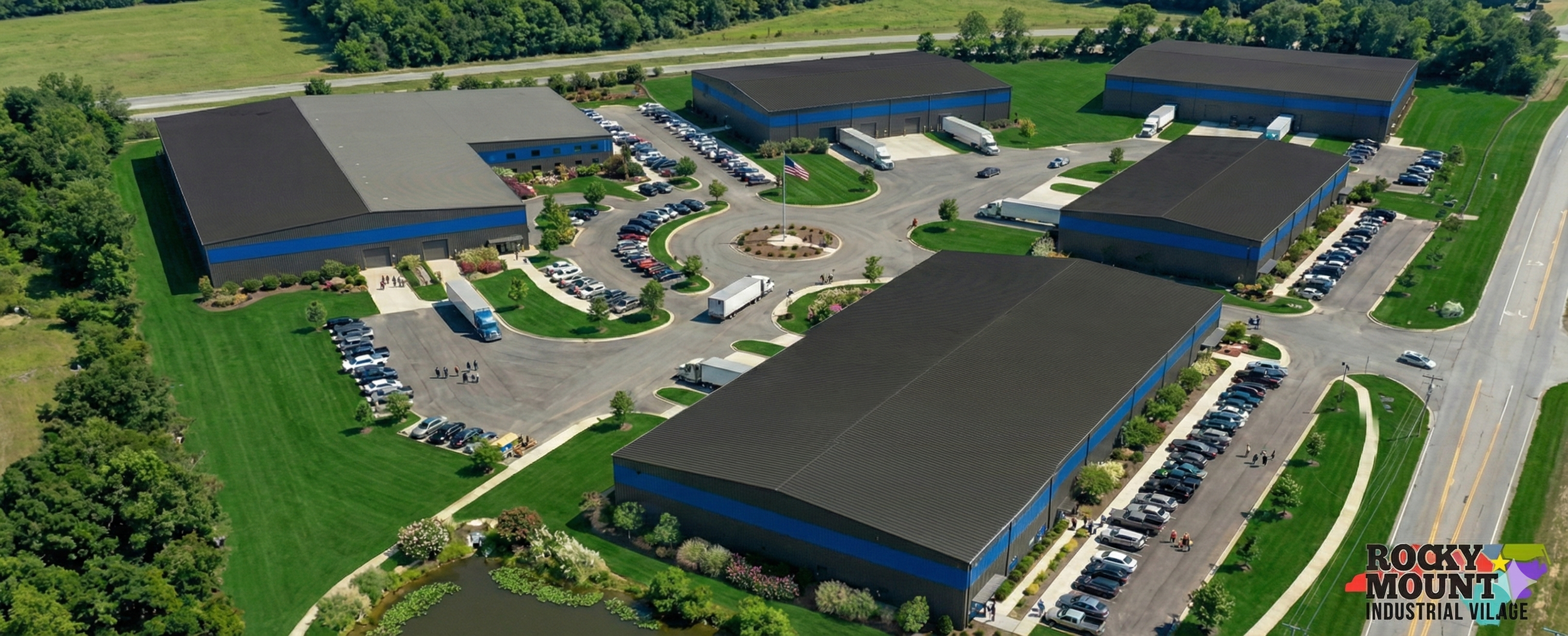 Aerial view of a commercial complex with large buildings, parking lots, trucks, and a central roundabout with a flagpole, surrounded by green lawns and trees.