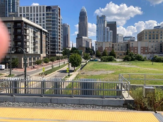 City skyline with modern high-rise buildings against a blue sky with clouds, viewed over a park area with grass and trees, and a street with crosswalks and sidewalks in the foreground.
