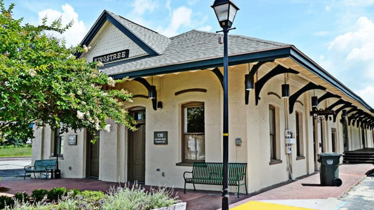 A small train station building with beige walls, black trim, and a gray shingle roof, featuring benches outside and a street lamp in front, under a blue sky with clouds.