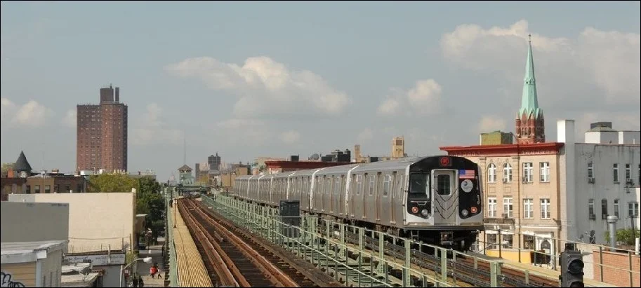 A train traveling on elevated tracks through an urban area with buildings and a church steeple in the background.