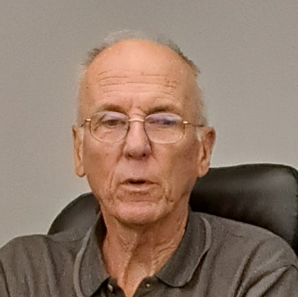 An elderly man with glasses sitting in a black office chair, wearing a gray polo shirt, against a plain gray background.