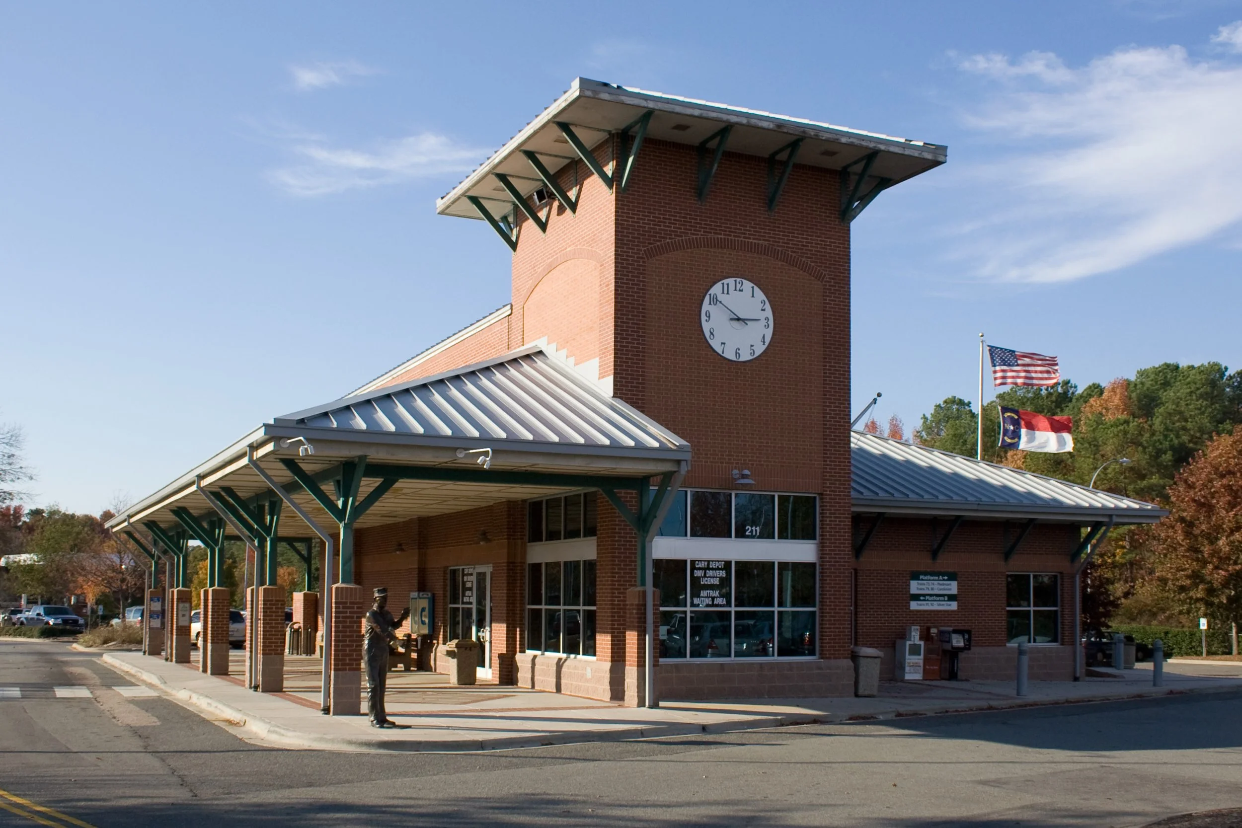 A brick train station with a large clock tower, American flags, and a person at a phone booth outside.