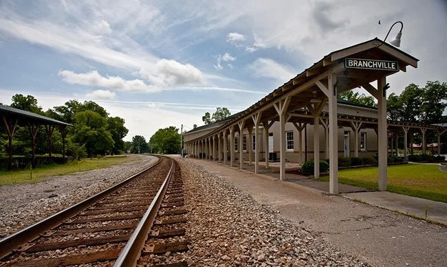 A vintage train station with a wooden platform and sign reading 'Branchville', beside railroad tracks and green trees under a partly cloudy sky.