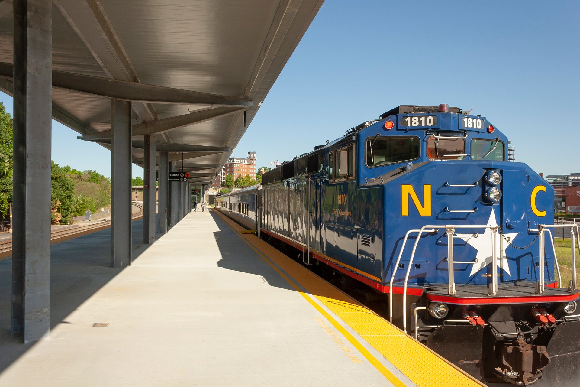 A blue locomotive train with yellow and white markings at a train station platform under a metal canopy, with a few people and buildings in the background.