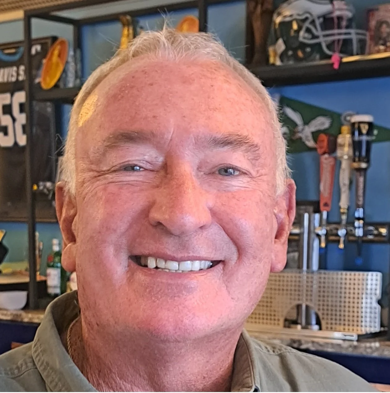 A smiling man with short, graying hair inside a sports-themed bar or restaurant, with sports memorabilia and beer tap handles in the background.