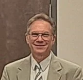 A man with glasses and gray hair, wearing a light-colored suit and a white shirt, standing indoors.