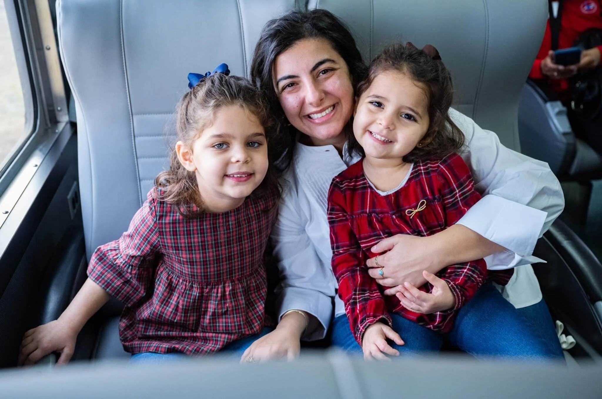 A woman with two young girls sitting together on a bus, all smiling at the camera. The women is in the middle, and the girls are on either side, dressed in red checkered tops.