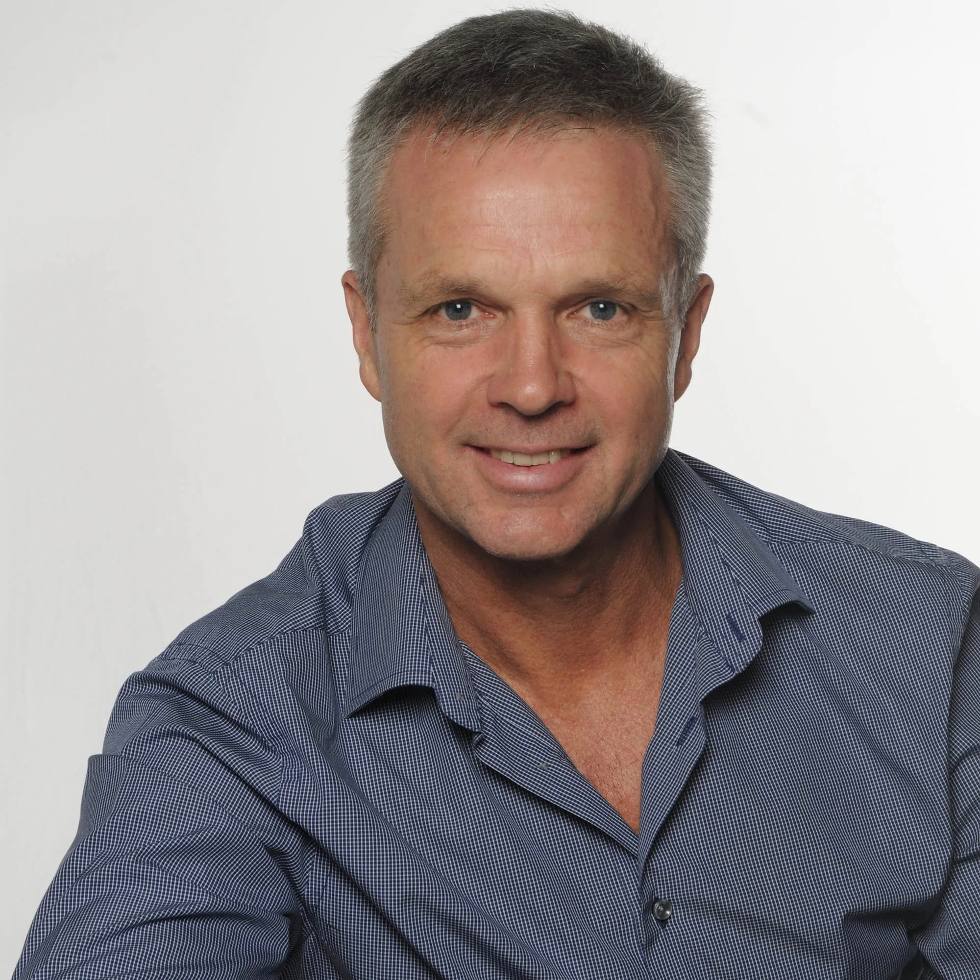 Portrait of a middle-aged man with short gray hair, blue eyes, wearing a blue patterned dress shirt, smiling against a plain white background.