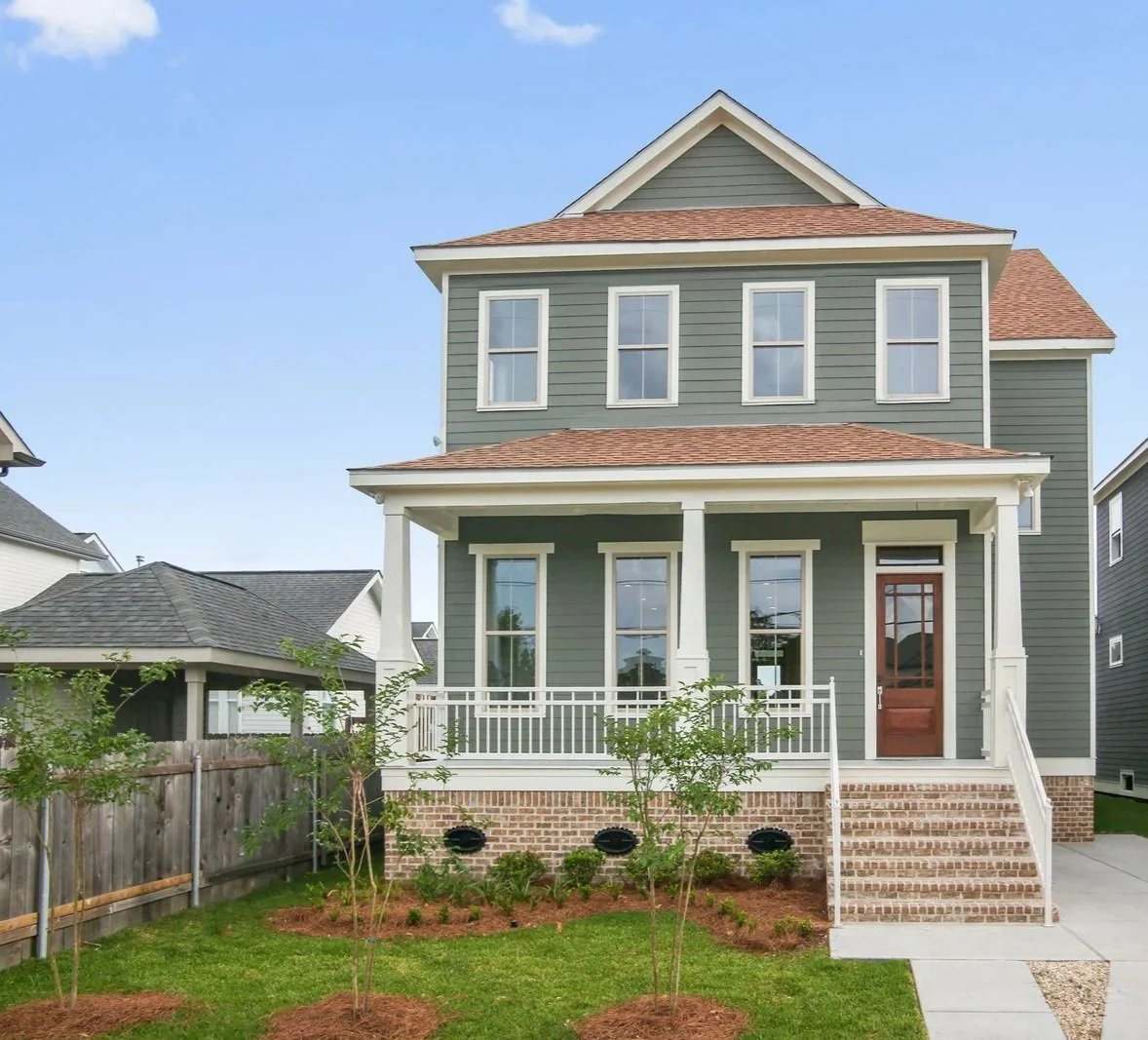 Front view of a two-story Southern Modern Farmhouse with green siding, a brick foundation, and a front porch with stairs, surrounded by a lawn and small trees.