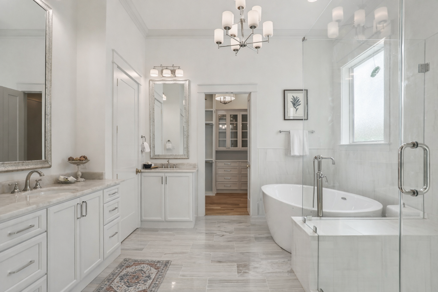 A spacious, modern bathroom with white cabinetry, marble countertops, large mirrors, a freestanding bathtub, a glass-enclosed shower, a window, wall art, and a chandelier light fixture.
