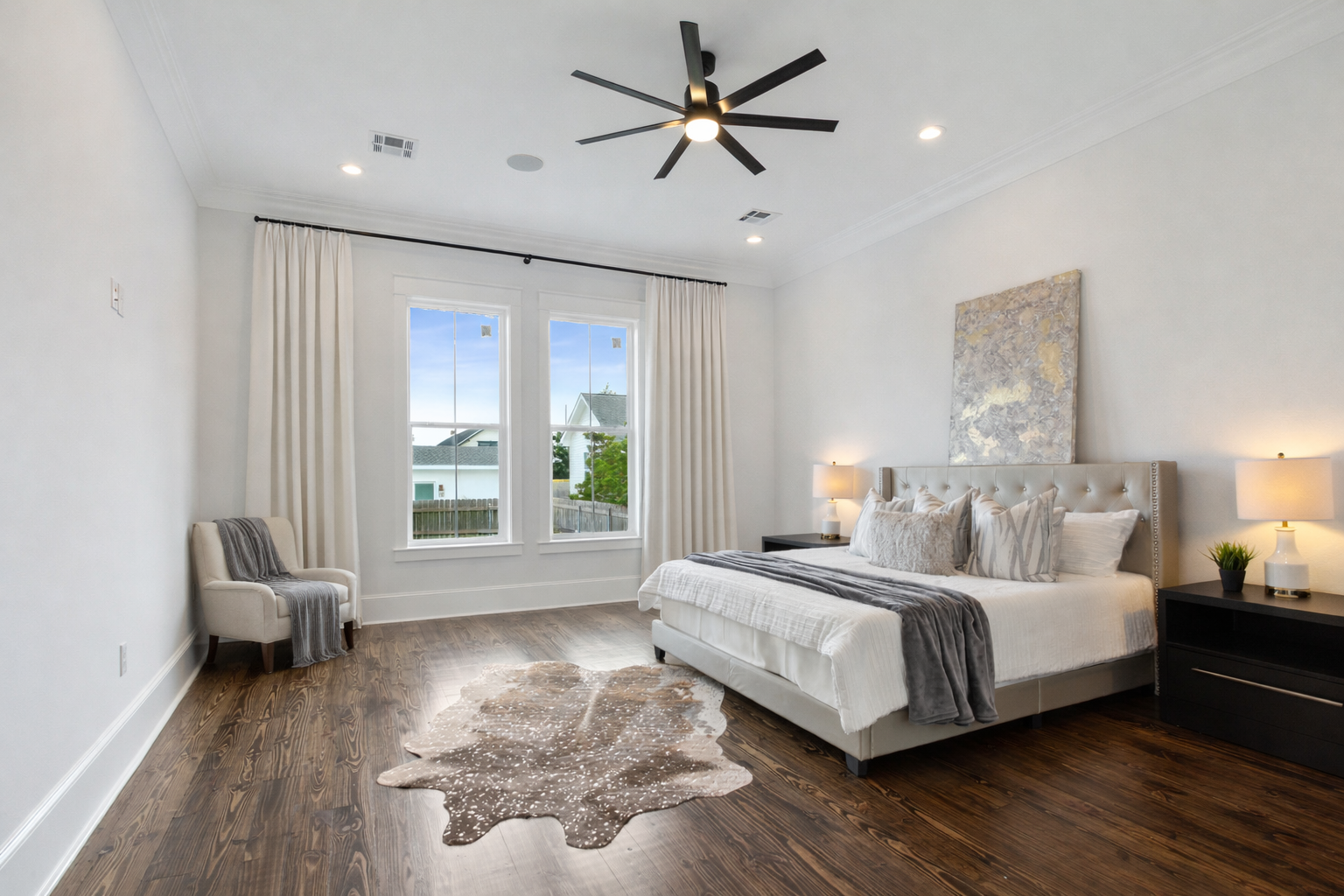 Master bedroom with walls in a beautiful shade of white, hardwood floors, a King sized bed with full bedding, nightstands with lamps, and a cowhide rug, large windows with linen drapes, a ceiling fan, and a piece of abstract artwork above the bed.