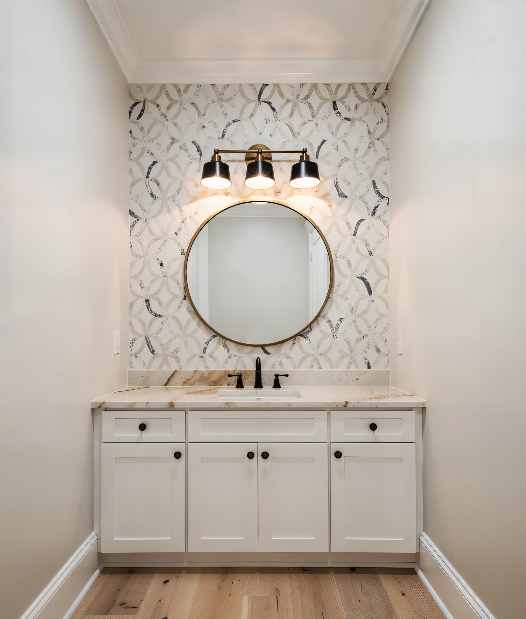 Bathroom vanity with white cabinet, marble countertop, round mirror, black faucet, decorative patterned wallpaper, and three black light fixtures above the mirror.