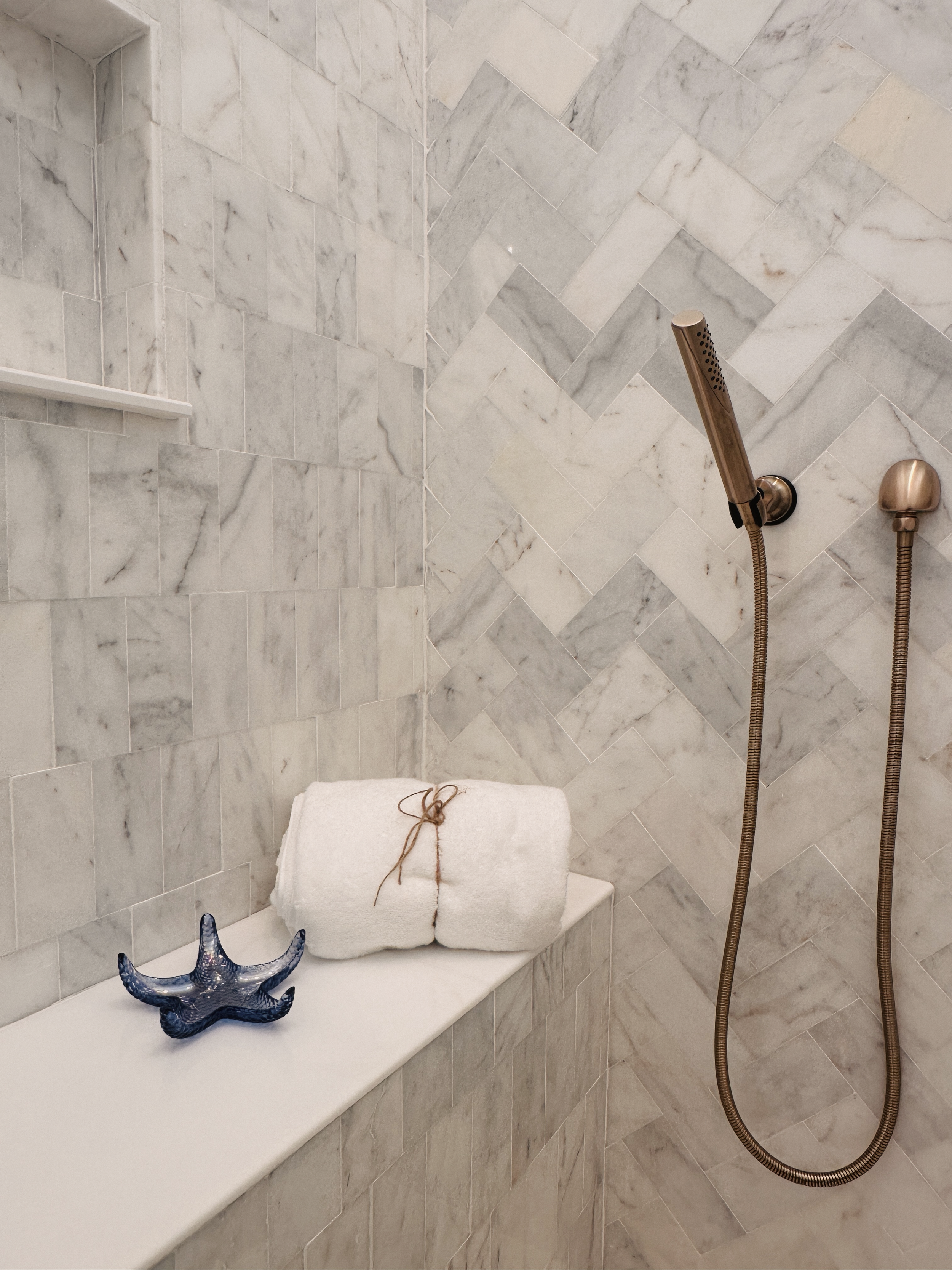 Marble-tiled shower with a rolled white towel tied with twine on a ledge, a blue starfish ornament, and a bronze handheld showerhead with a wall-mounted support.
