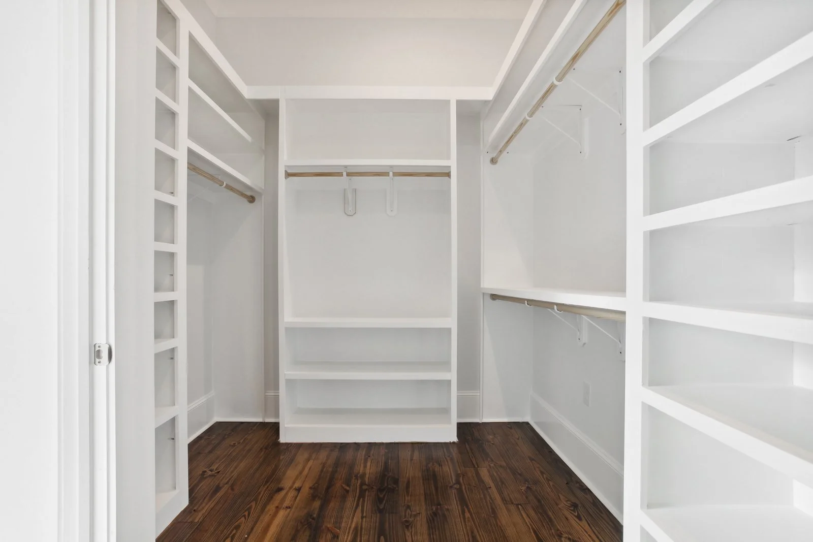 Empty walk-in Master closet with white shelving, rods for hanging clothes, and dark wooden floor.