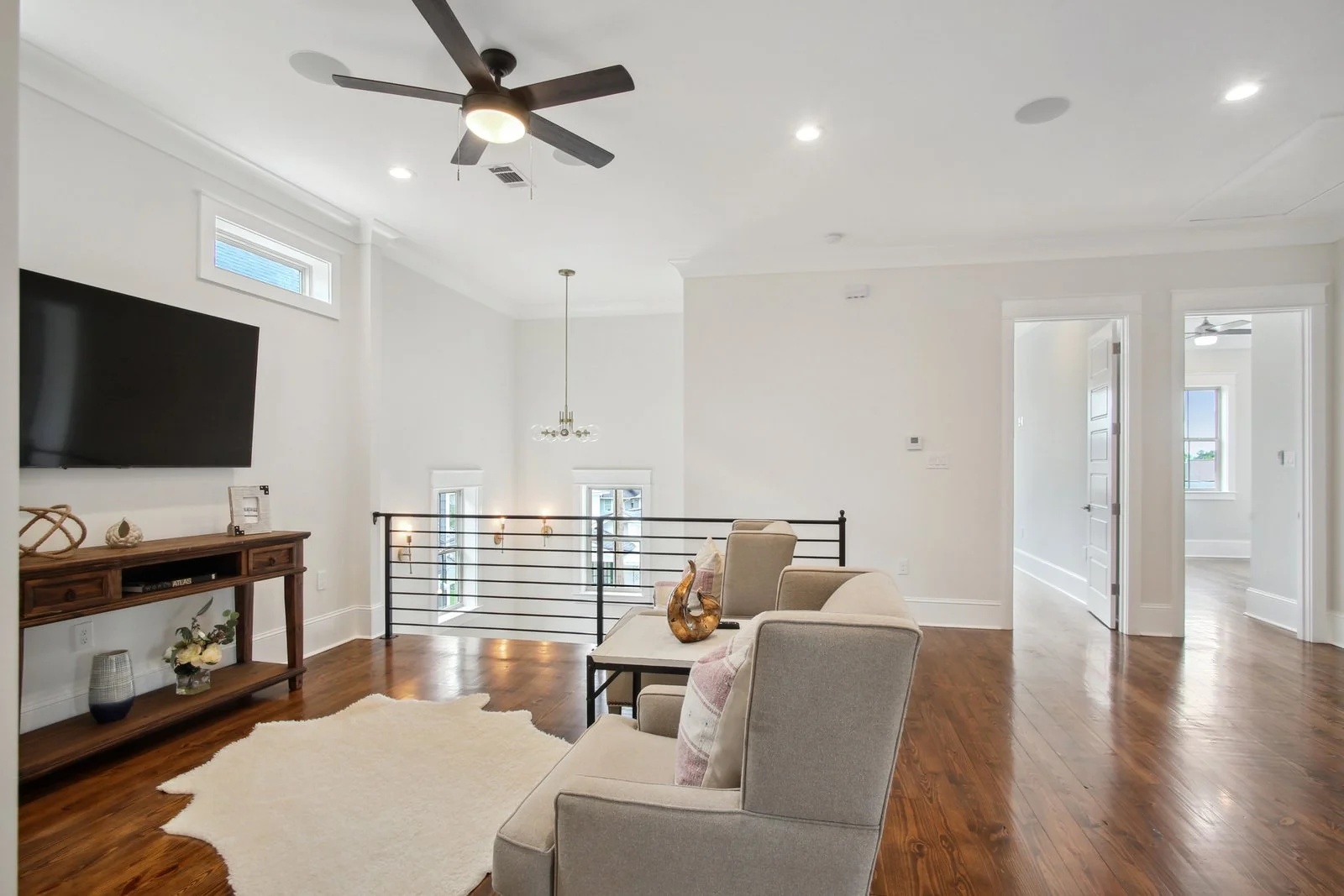 A modern upstairs den with white walls, hardwood floors, high ceilings, a ceiling fan, a wall-mounted TV, beige seating, a small side table, and minimal decor.