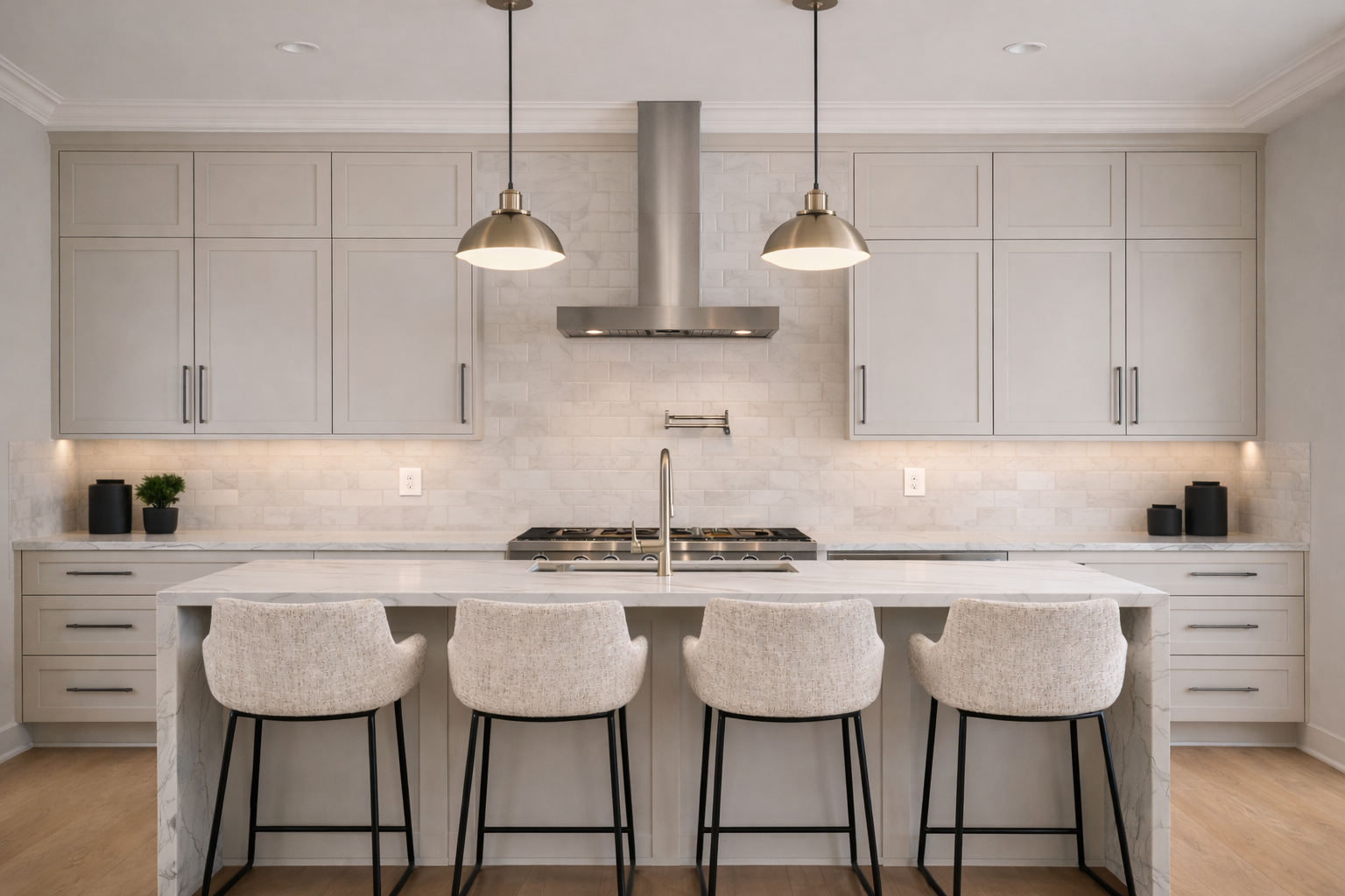 Modern kitchen with light gray cabinets, a marble island with beige upholstered chairs, stainless steel range hood, black pendant lights, and minimalist decor.