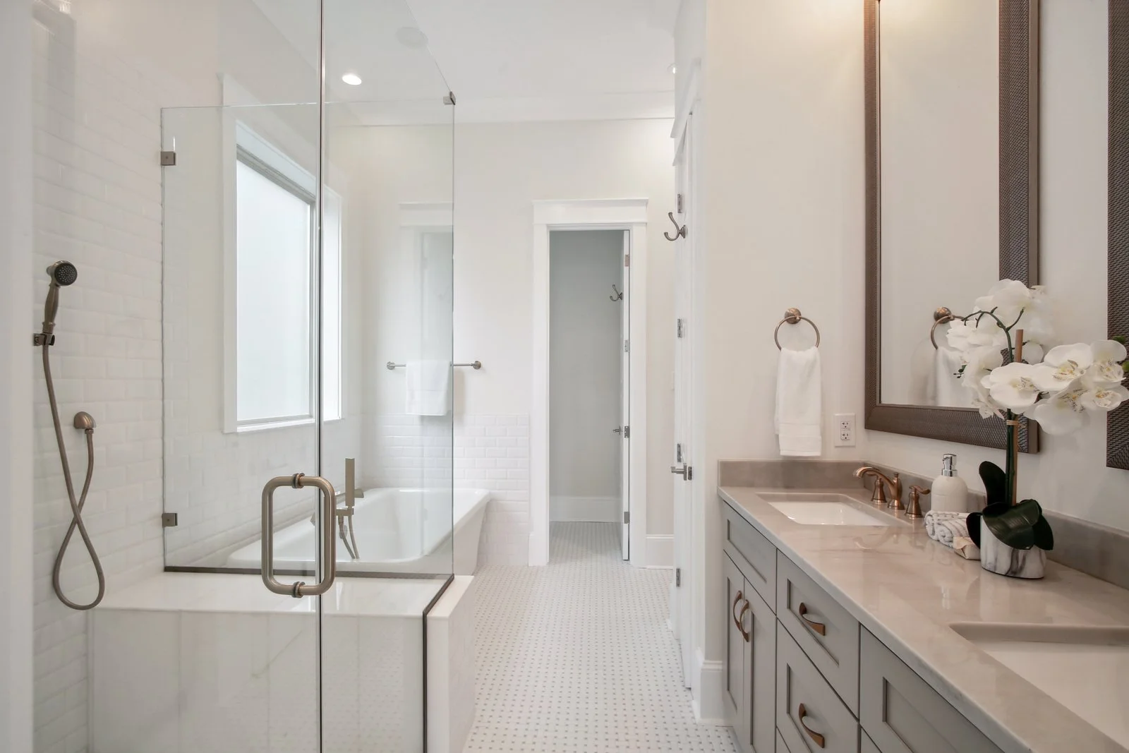 Modern primary bathroom with glass-enclosed shower, free-standing bathtub, dove gray vanity with cream stone counter, beautiful gray and white mosaic floor tiles, large mirror, and decorative orchids.
