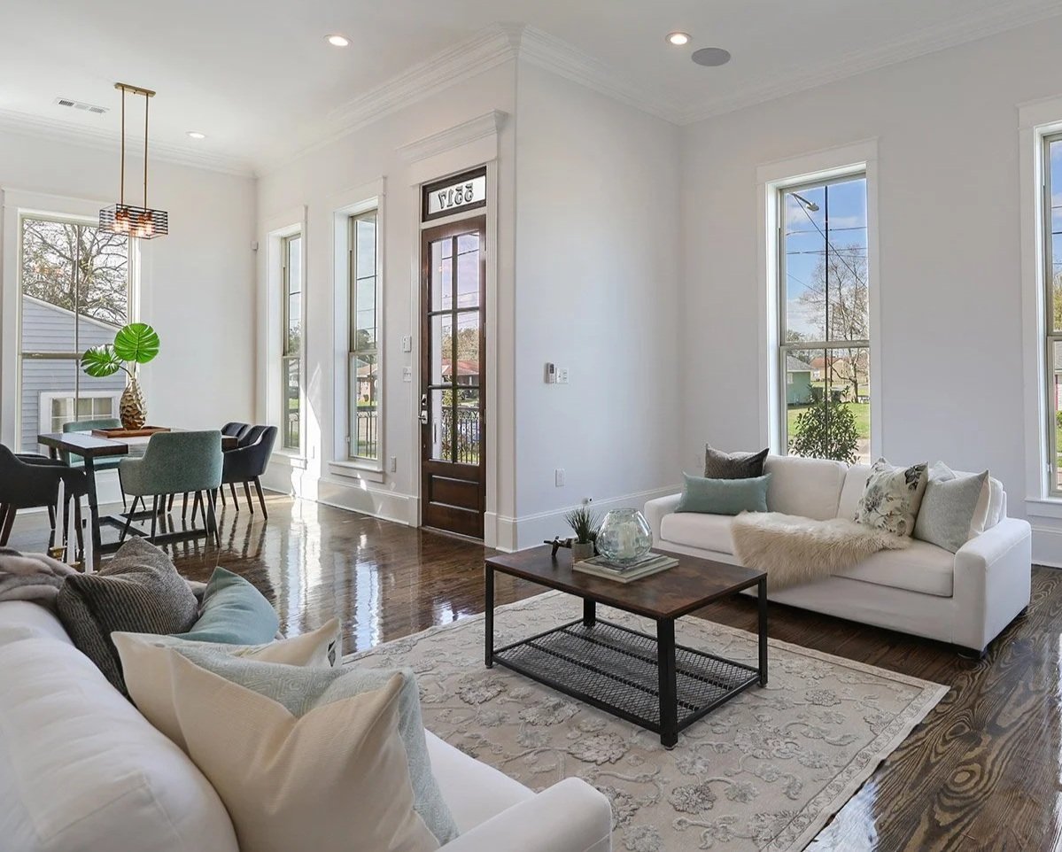 Bright living room with white walls, large windows, wooden floors, a white sofa with cushions, a coffee table with décor, and a dining area with a table and chairs, illuminated by natural light.