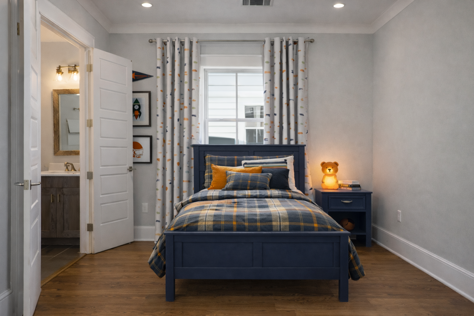 A bedroom for a little boy with a blue bed, plaid bedding, colorful pillows, and a nightstand with a bear lamp and books. There are curtains, wall art, and an adjacent bathroom visible.