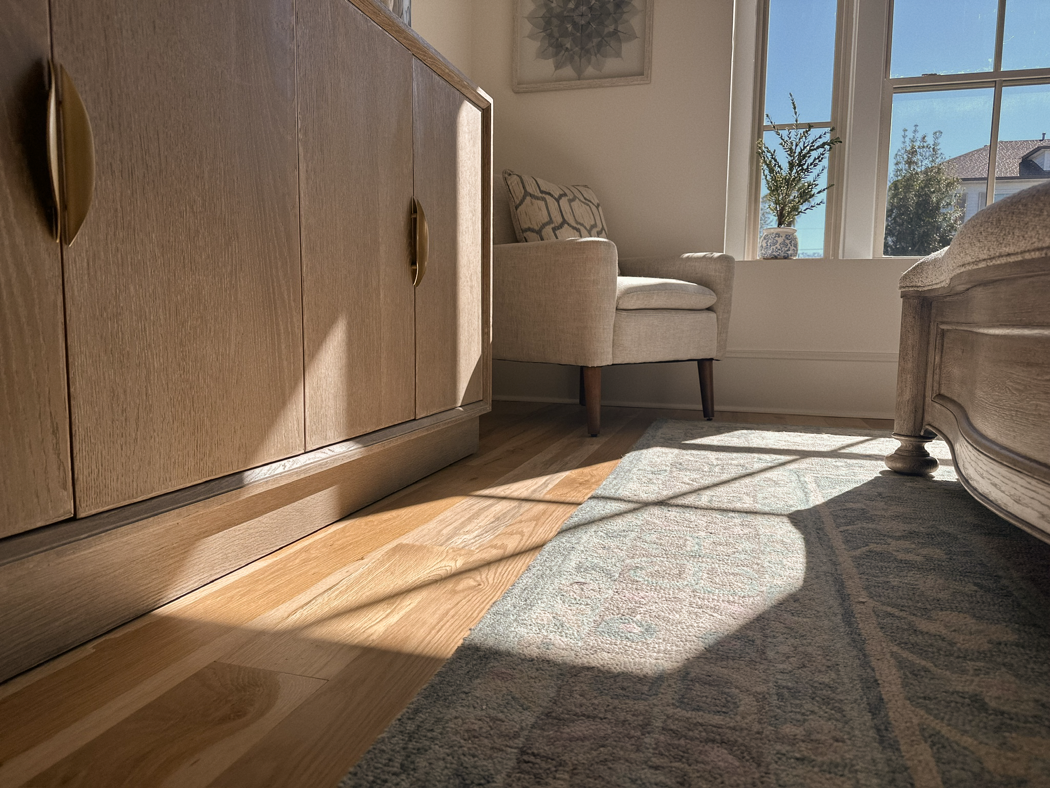 Sunlit bedroom corner with a wooden dresser, beige armchair, decorative pillow, window with plants, and a partially visible wooden bed frame with a rug on the floor.