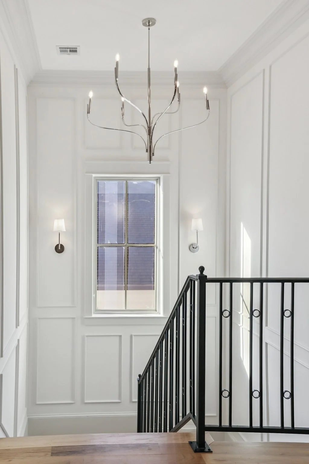 A staircase landing with white paneled walls, a window with blinds, black metal railing, and a modern chandelier with multiple lights hanging from the ceiling.