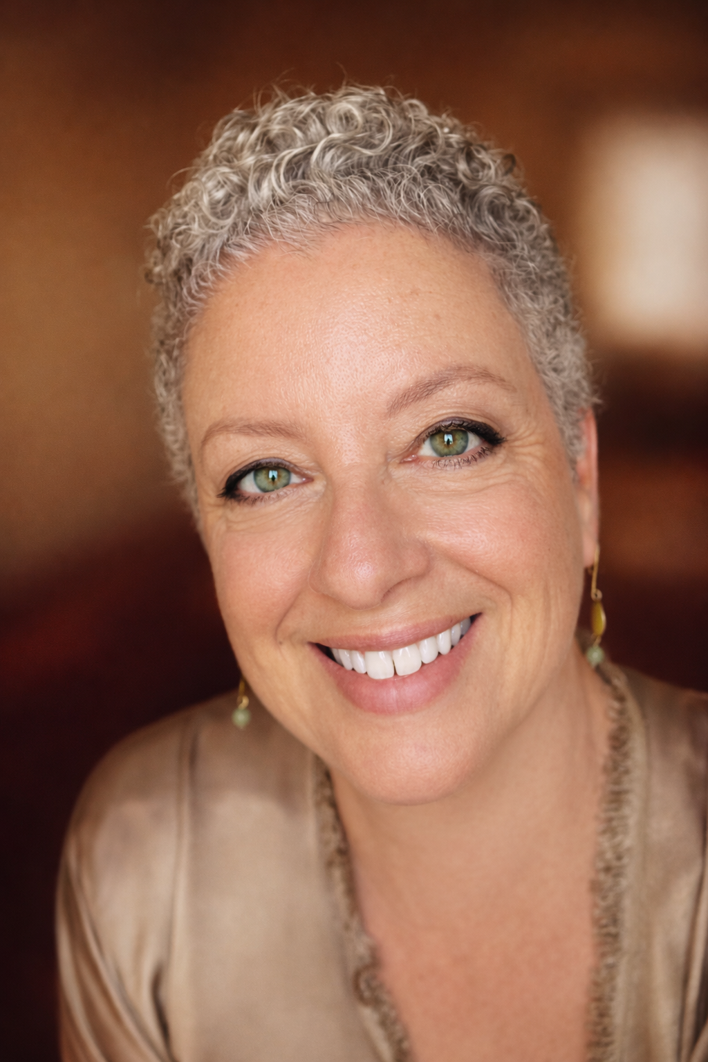 Close-up portrait of a smiling woman with short, curly gray hair and green eyes, wearing earrings and a beige top.
