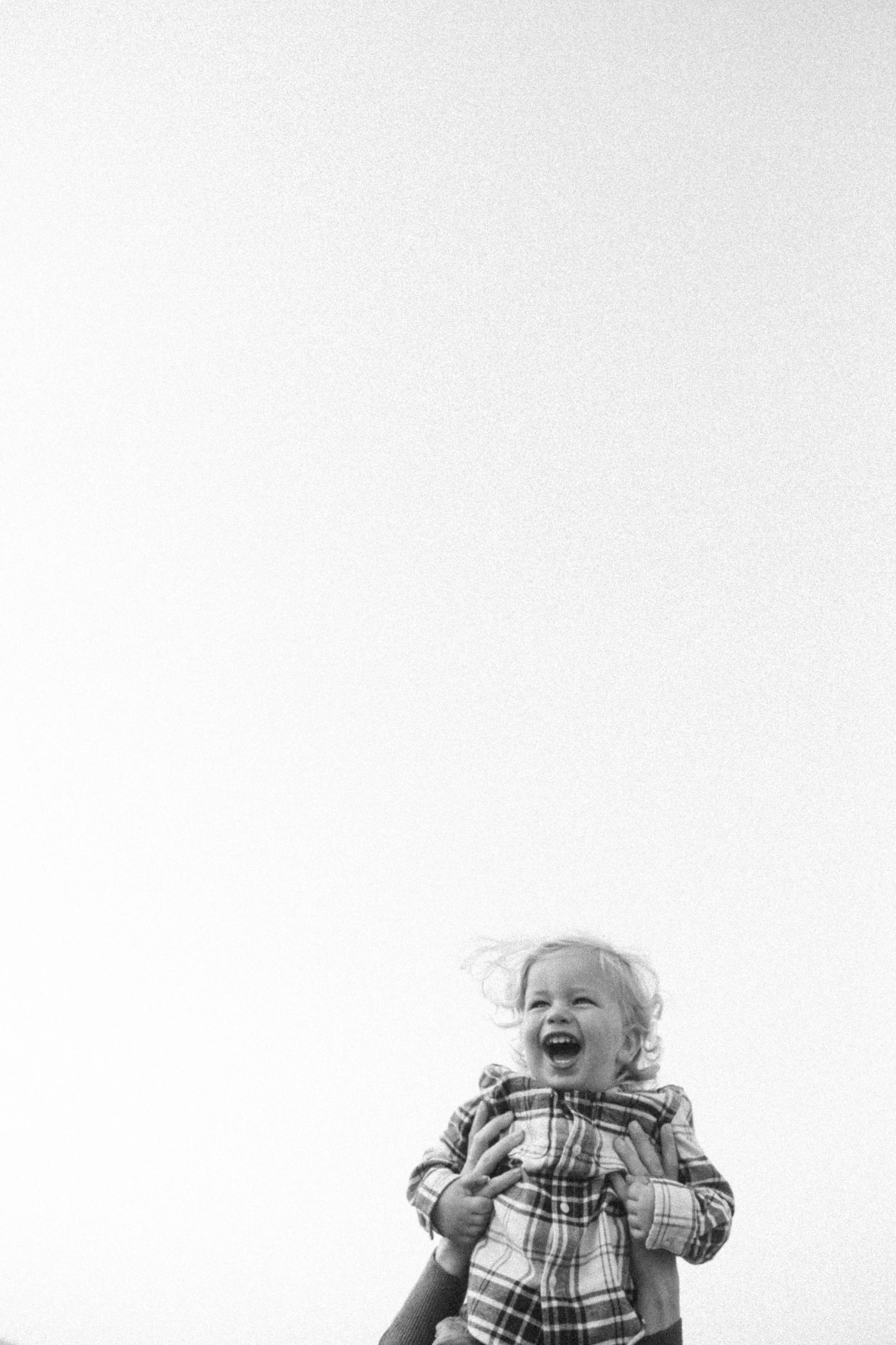 A young girl with curly hair laughing happily while being held up in the air outdoors against a plain sky background.