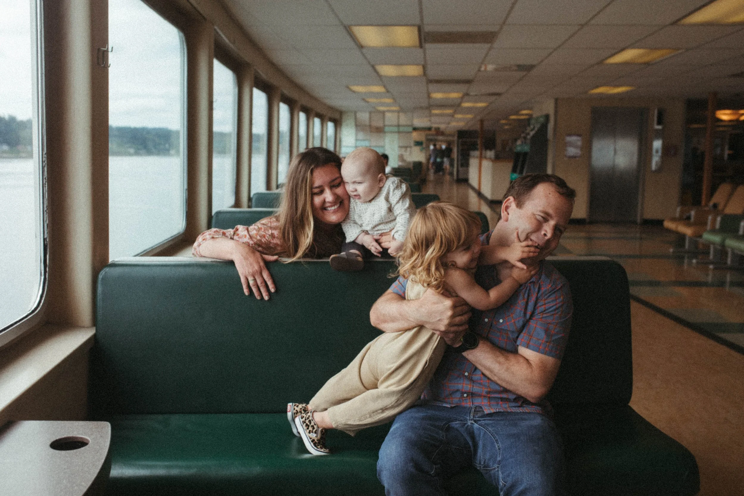 Family of four happily playing and teasing each other on a ferry, with large windows showing water outside.