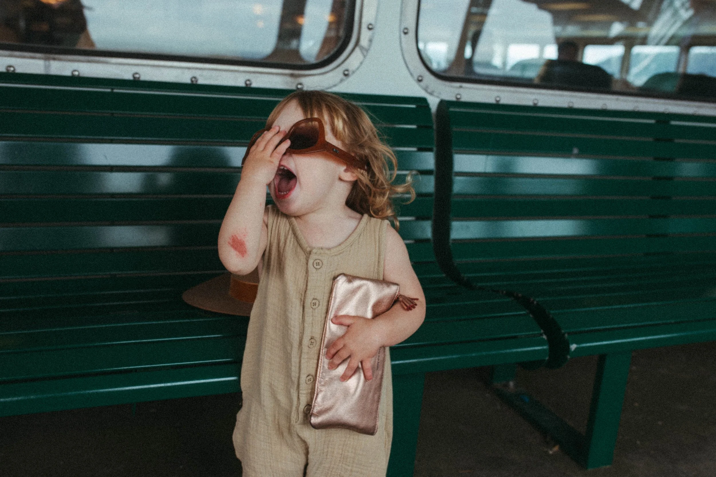 A young child with curly red hair wearing a beige outfit and sunglasses, standing on a green park bench in a train station with a high speed train in the background, holding a gold clutch bag, which is partially covered by her hand, with a red lipstick stain on her arm and her mouth open as if talking or singing.